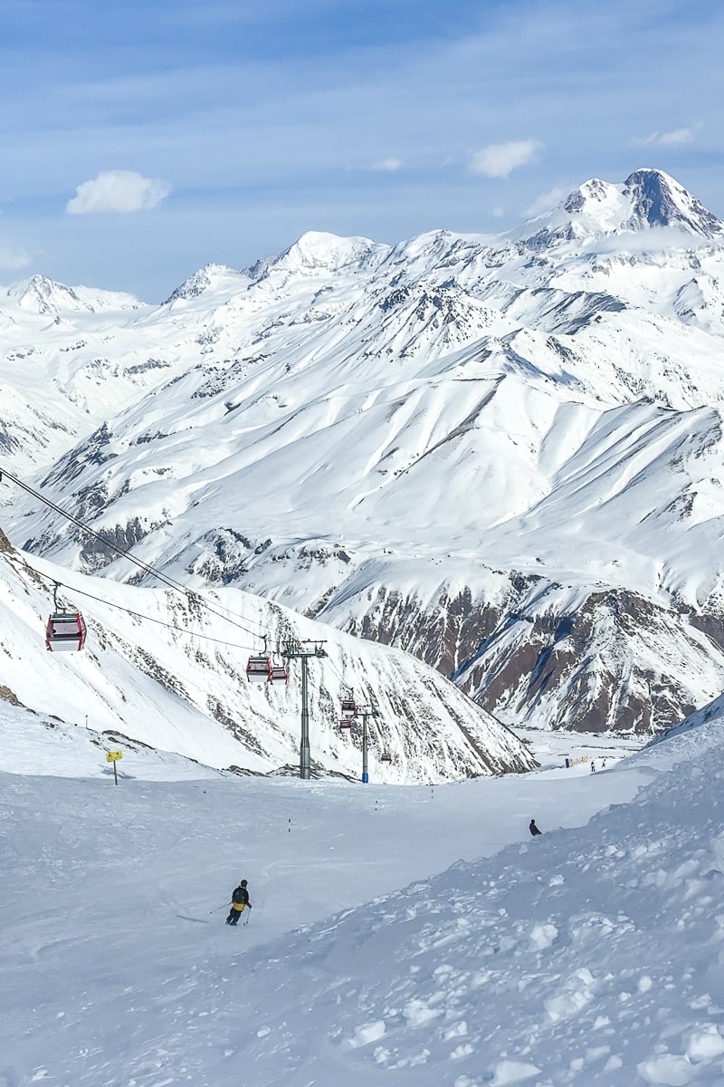 Two people skiing in the distance as a chairlift passes, surrounded by huge snowy, mountains