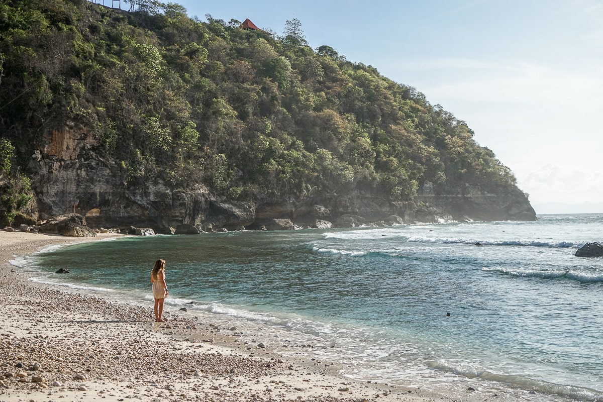 A woman stood on the water's edge looking out to the ocean, a tree covered cliff area in the background
