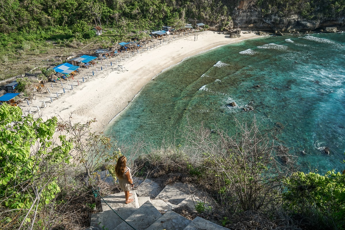 A woman stood on a high cliff looking down on a sandy beach and crystal blue waters