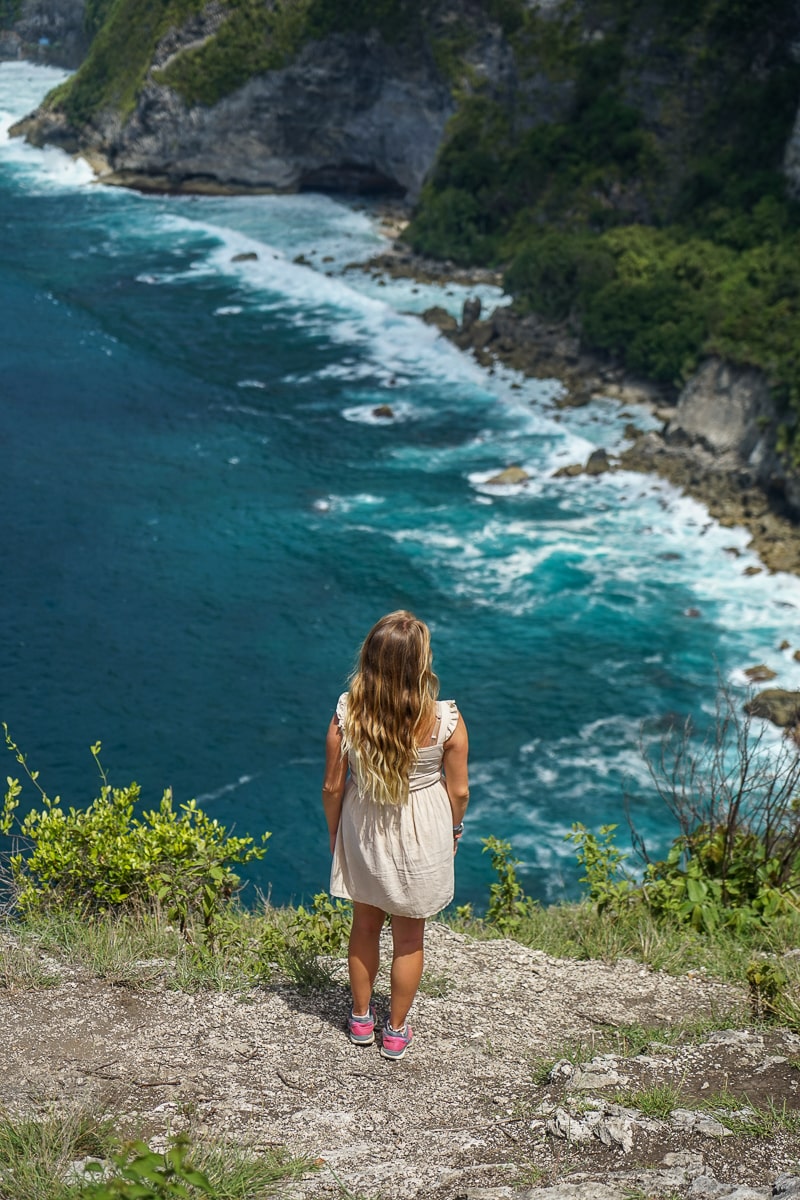 A woman stood on the edge of a cliff in a cream dress overlooking azure blue waters
