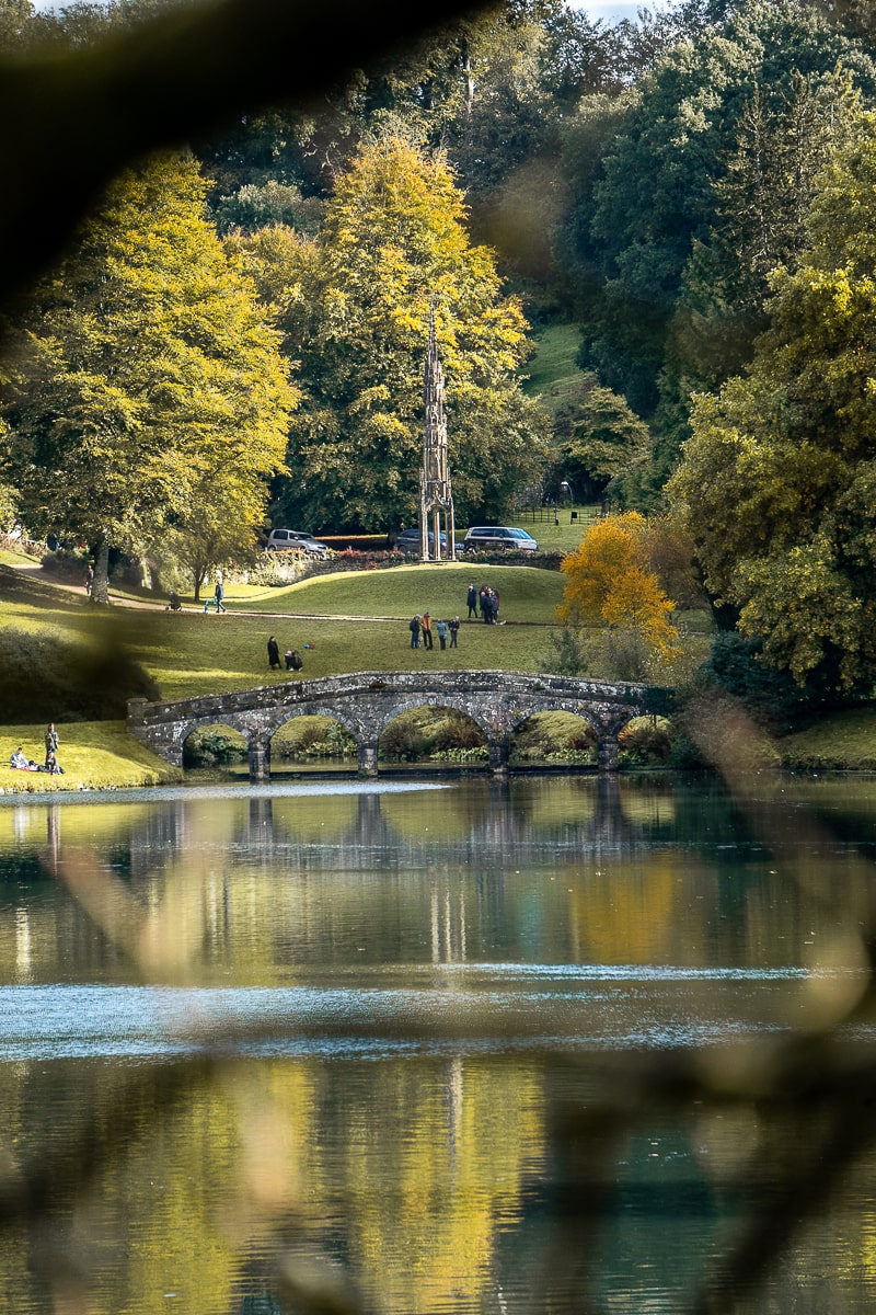 A palladian bridge across a calm lake with a gothic spire in the background