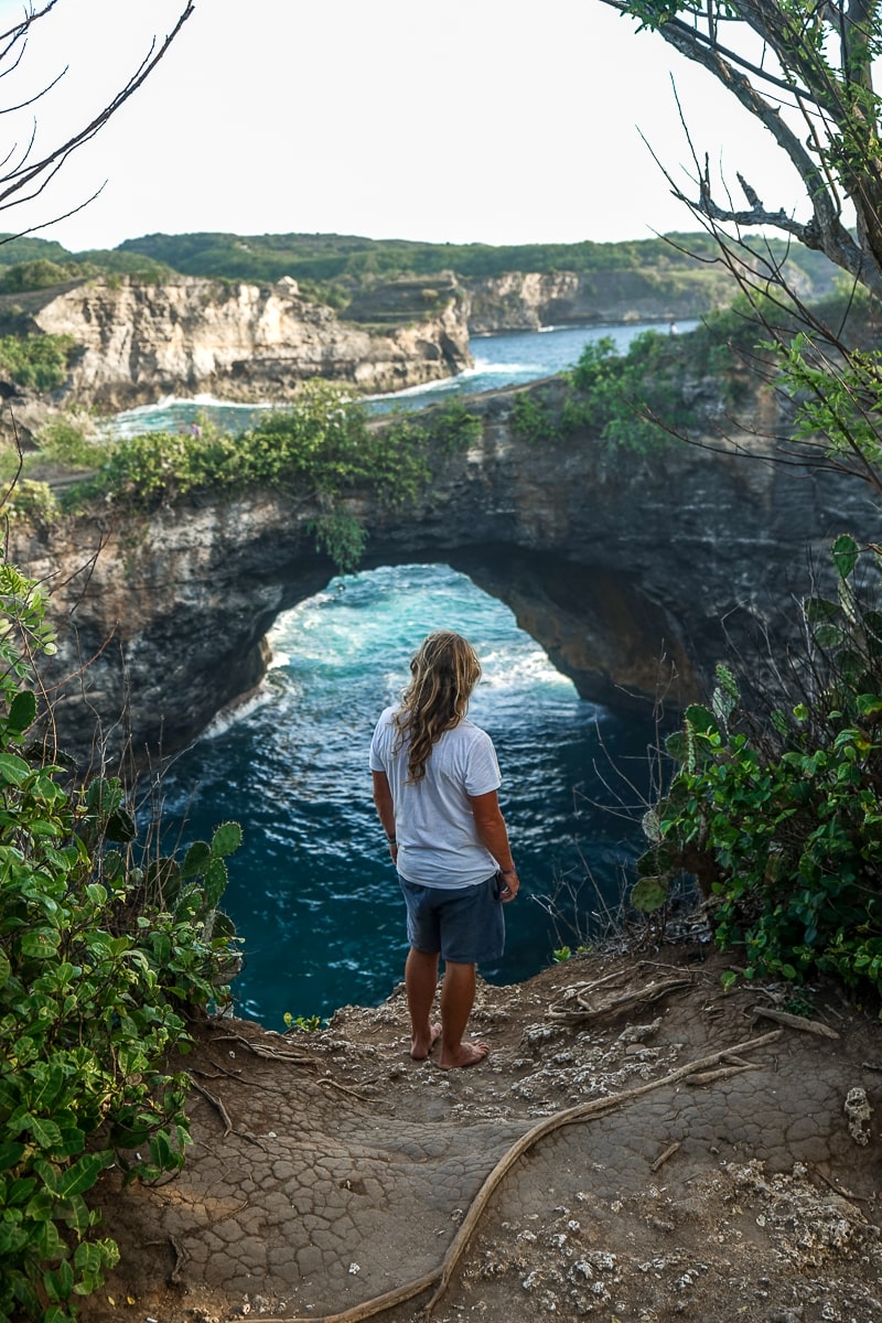 A man stood on the edge of a cliff with a natural arch in the rocks leading to a cove