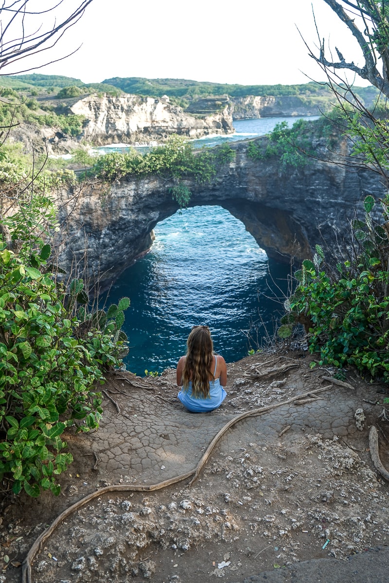 A woman sat on the edge of a cliff with a natural arch in the rocks leading to a cove