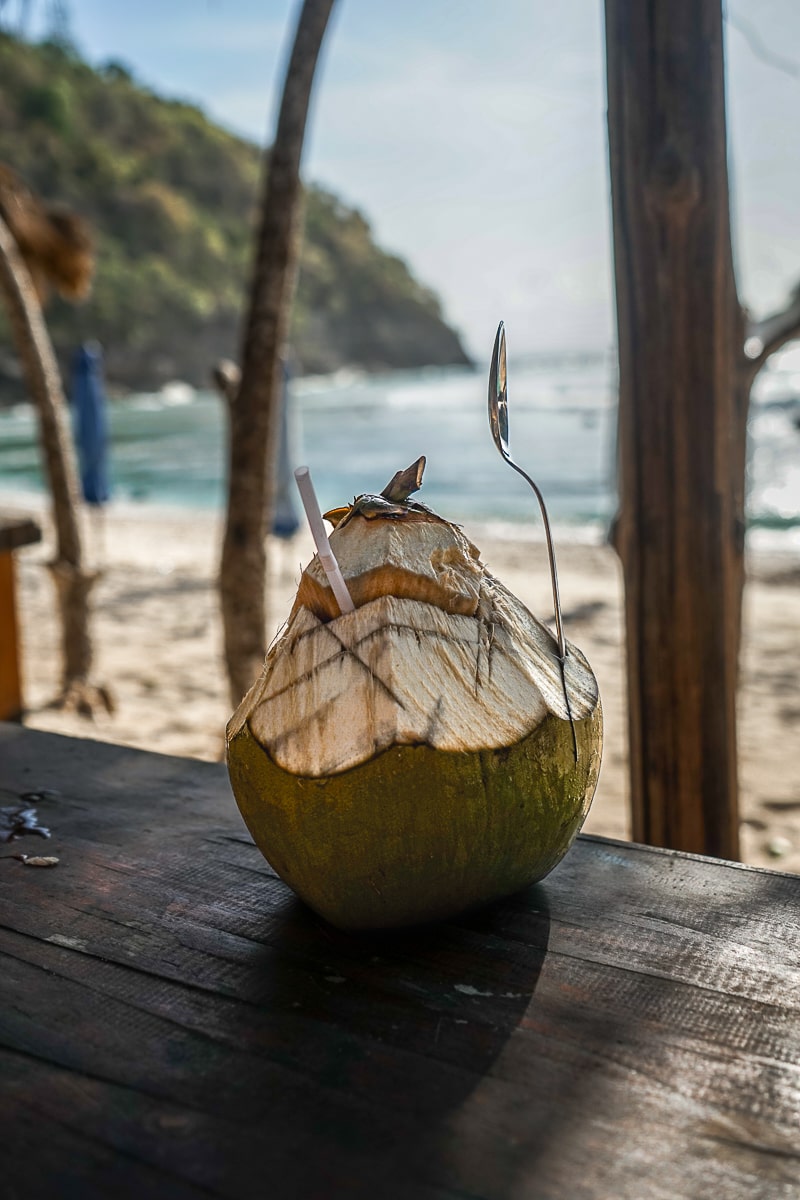A coconut with straw and spoon on a wooden table, the ocean in the background