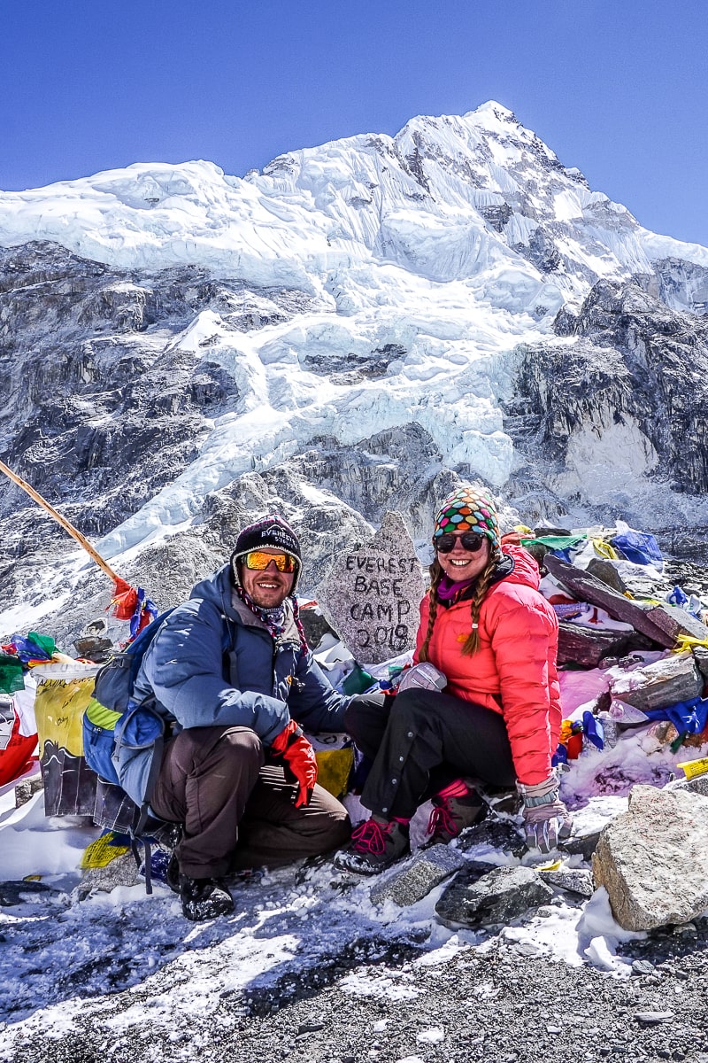A couple in winter clothes crouching near the "Everest Base Camp 2019" stone with a huge snowy mountain behind