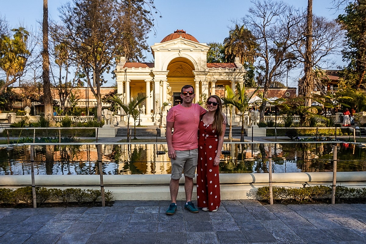 A couple stand arm in arm in a landscaped garden with a neo-classical building in the background