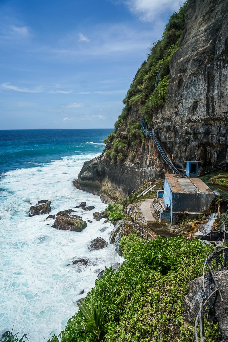 A steep rocky cliff towering over the white foamy ocean and a small blue shack