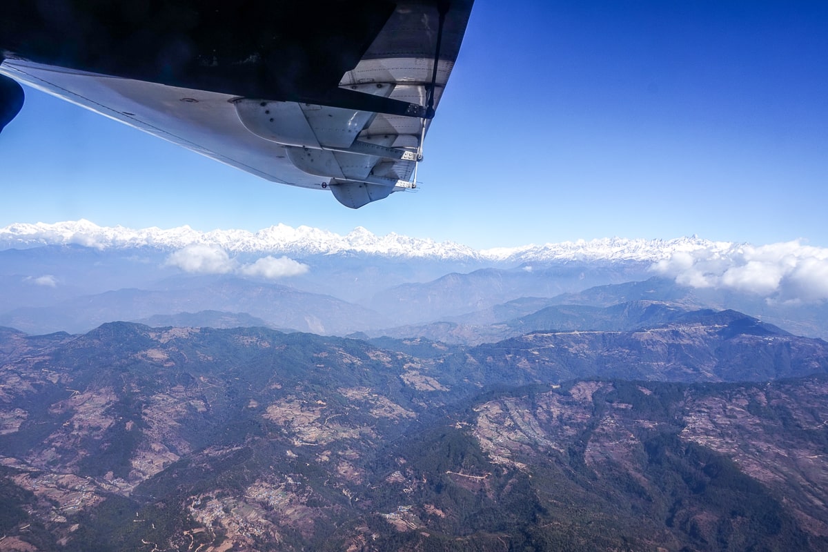 A wide landscape view of the snow-capped Himalayas from a plane. The plane's wing is visible in the top of the photo