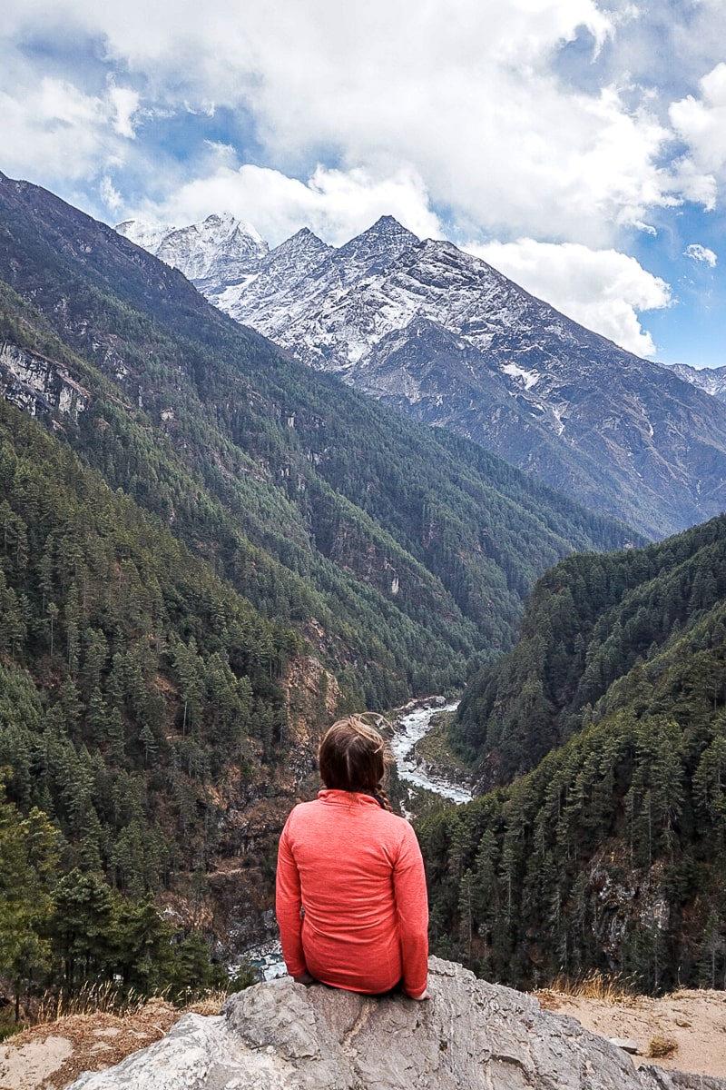 A woman in a pink jacket sat on a rock overlooking a deep ravine and river. A snow capped mountain towers in the distance