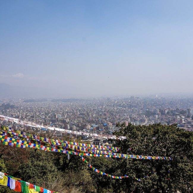 A panoramic view of Kathmandu, densely packed houses stretching to the horizon. Strings of prayer flags hang in the foreground
