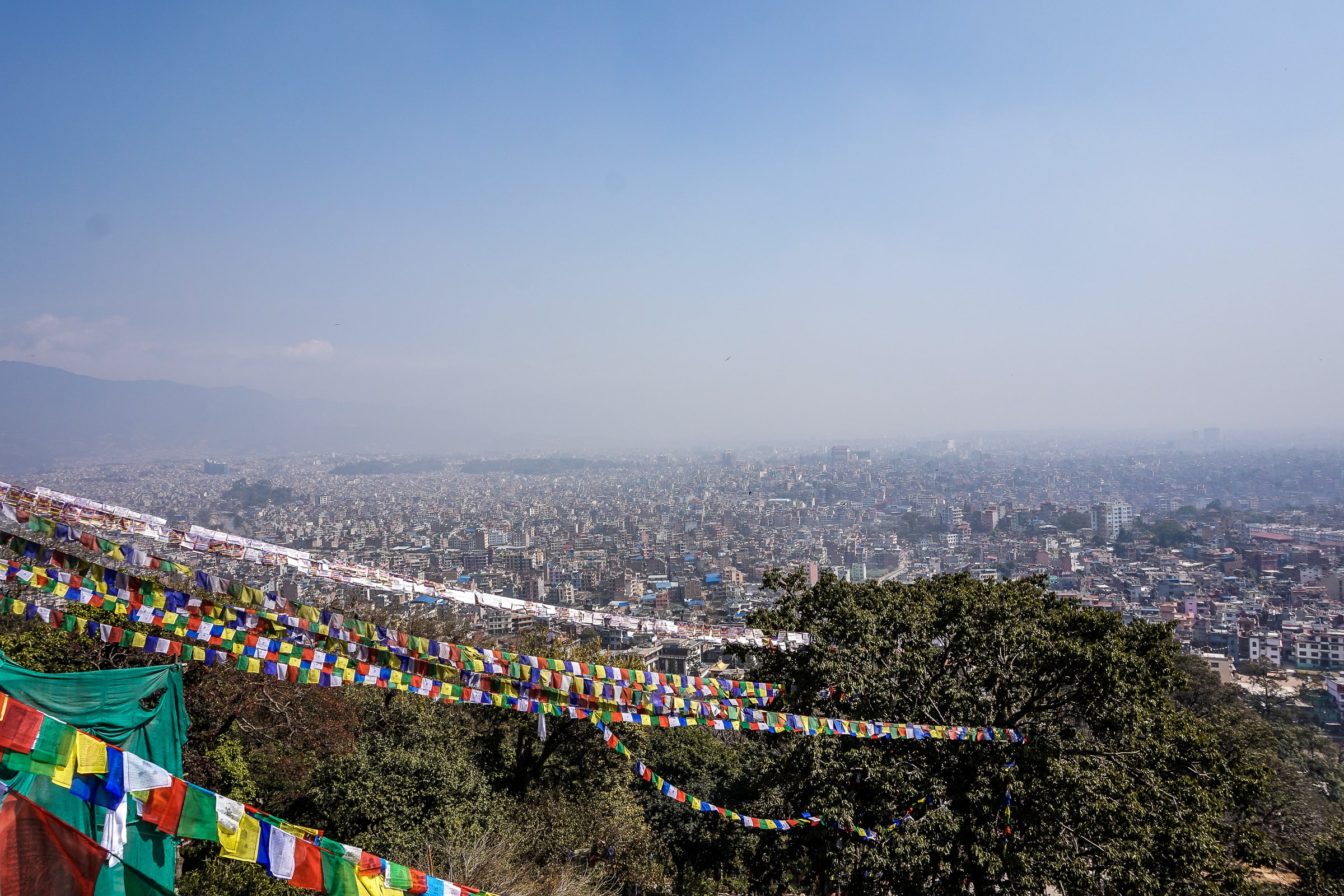 A panoramic view of Kathmandu, densely packed houses stretching to the horizon. Strings of prayer flags hang in the foreground