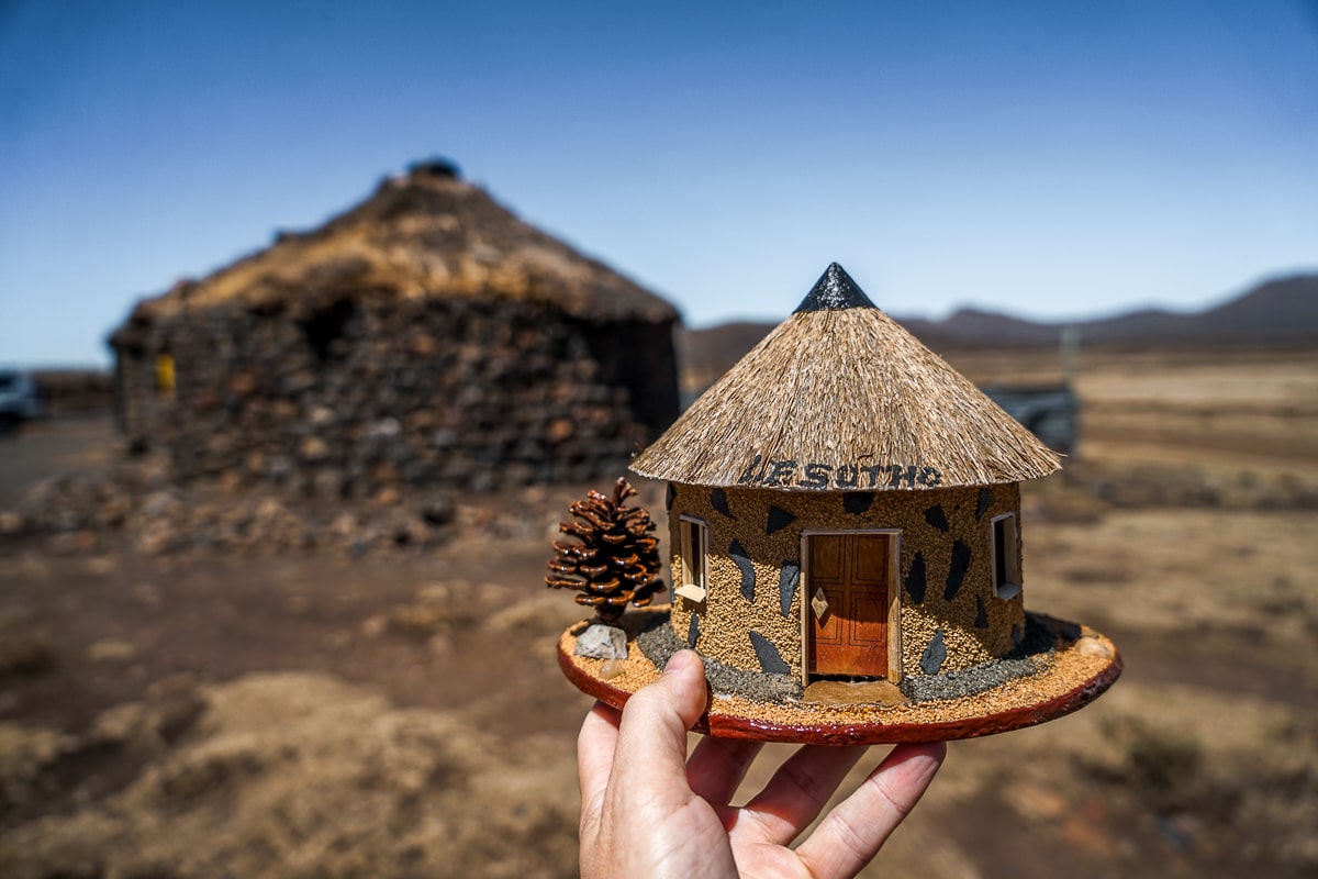 A man holding a miniature handcrafted sculpture of a Lesotho hut in front of a real hut