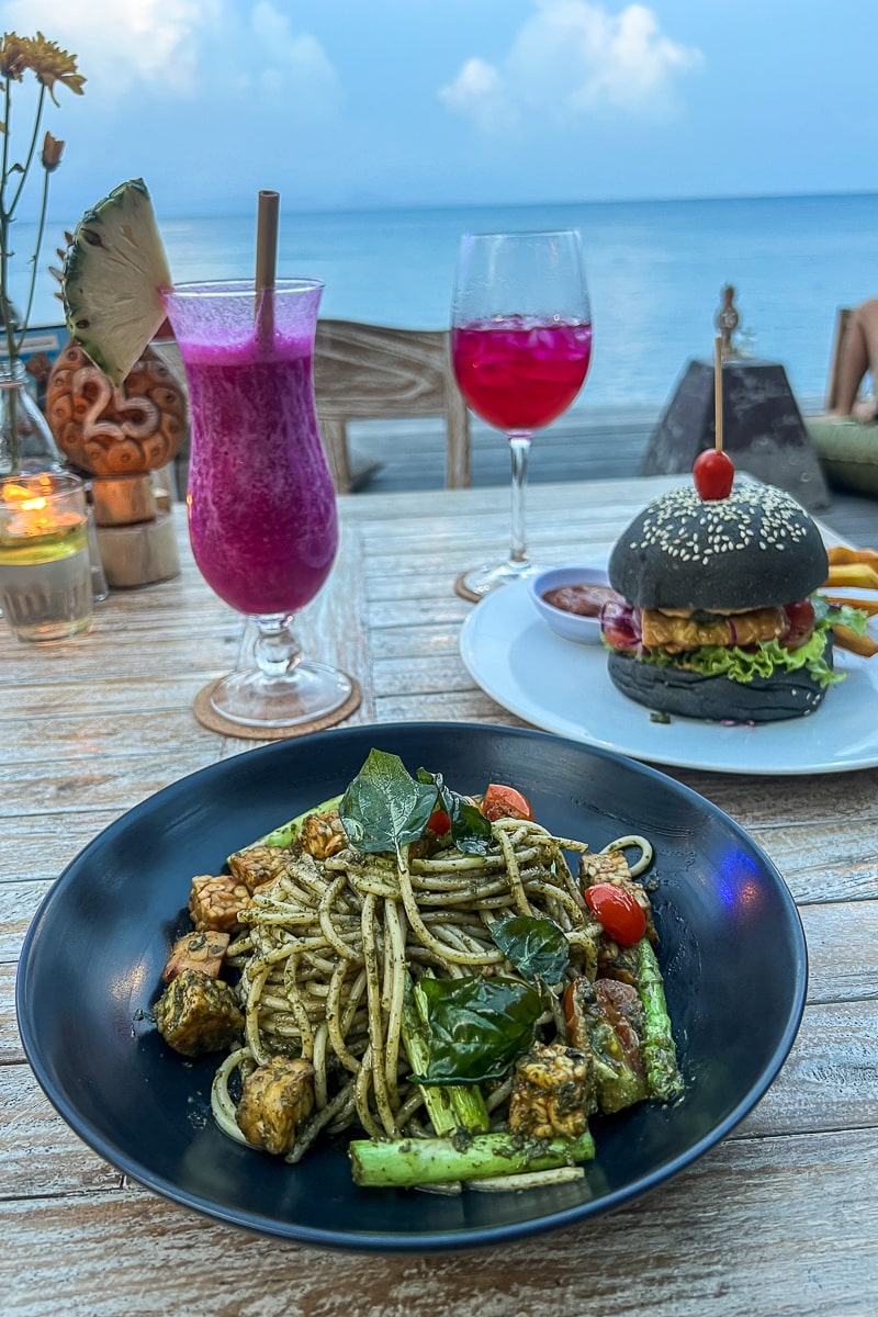 A pesto pasta dish and a burger with black bun and a couple of pink cocktails