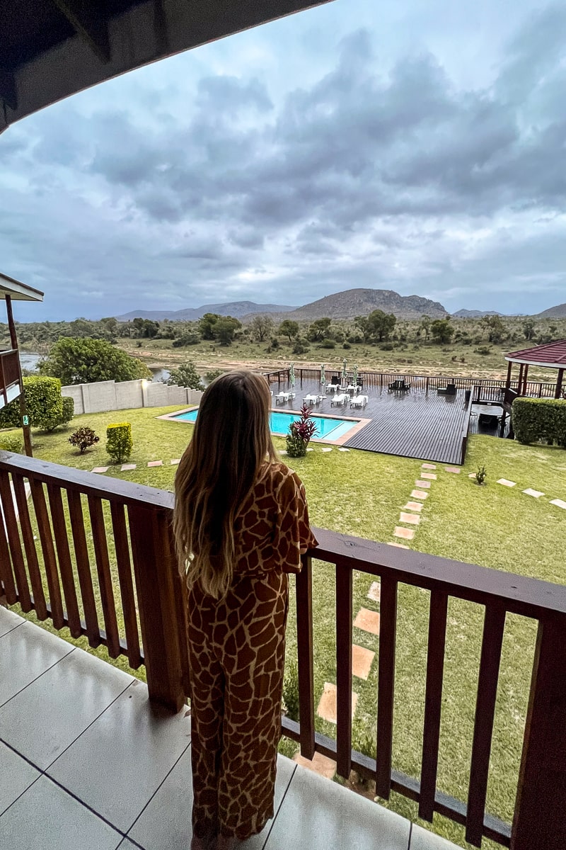 A woman leaning on a wooden balcony overlooking a pool and garden