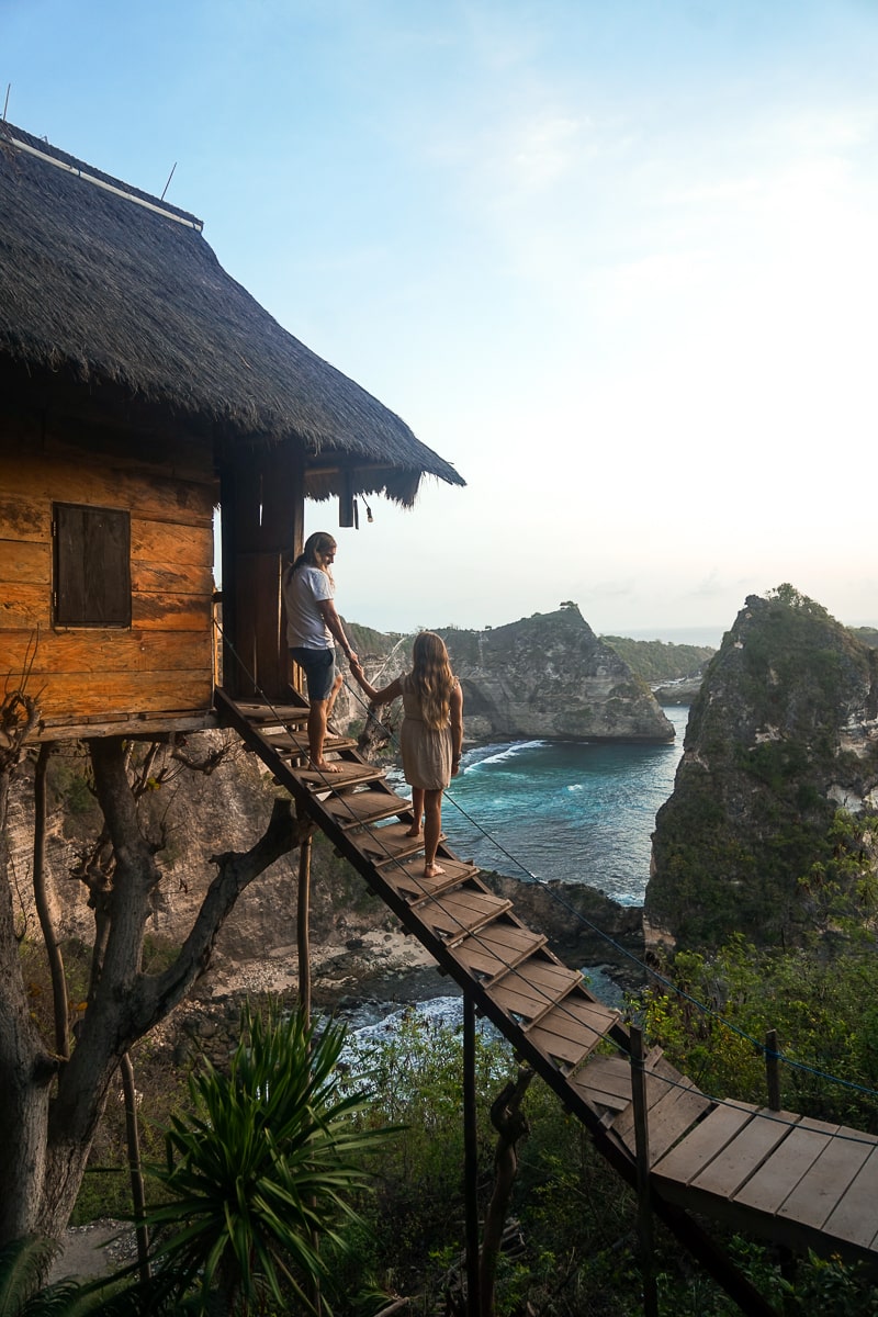 A couple standing hand-in-hand at the top of a set of stairs leading to a wooden tree house with a view of the ocean and islands