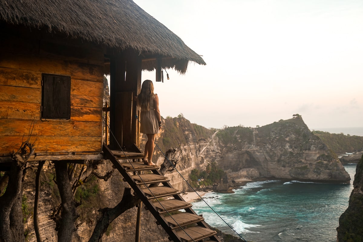 A woman standing at the top of a set of stairs leading to a wooden tree house with an incredible view of the ocean and islands