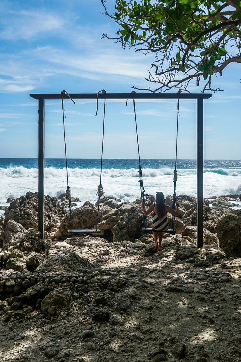 A set of two swings overlooking the ocean. A woman is sitting on the right swing.