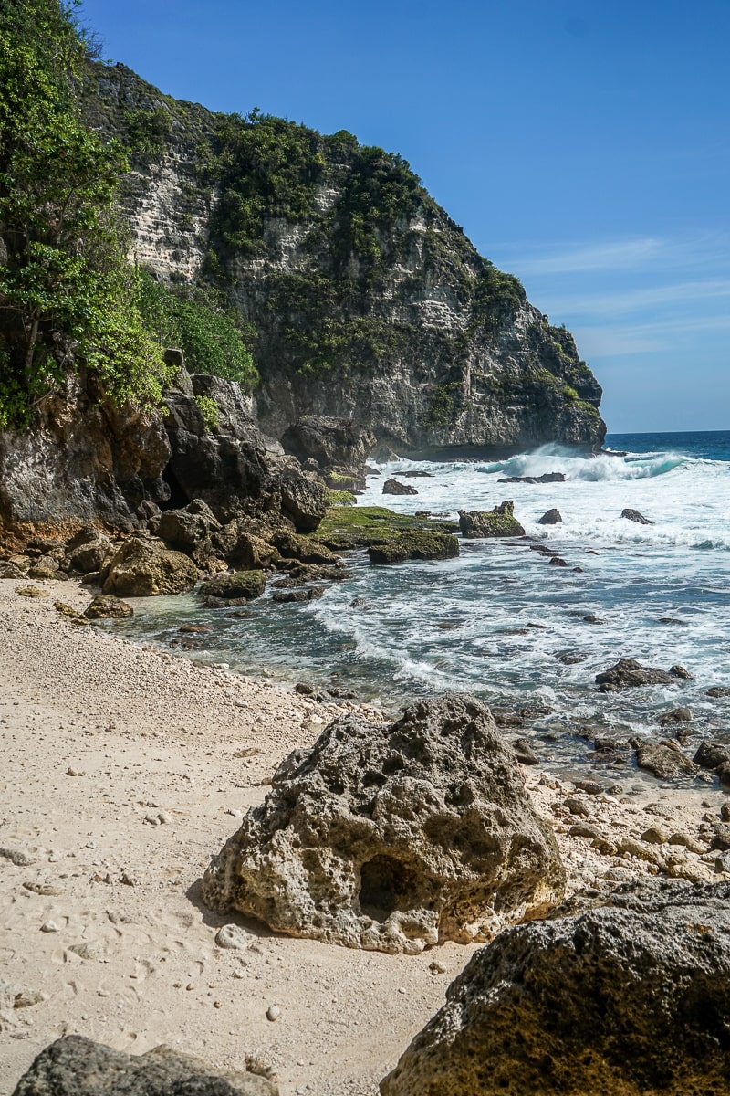 A sandy beach with rocky areas and white foam sea surrounded by cliffs