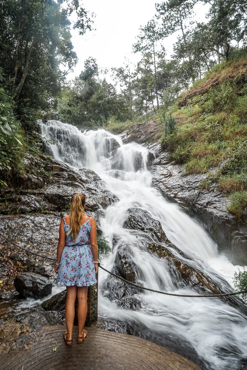 A woman standing in front of a cascading waterfall with lush vegetation on either side