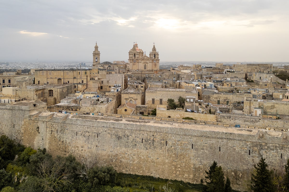 Aerial view of a walled city made with sand colored limestone. A cathedral prominently standing at the highest point
