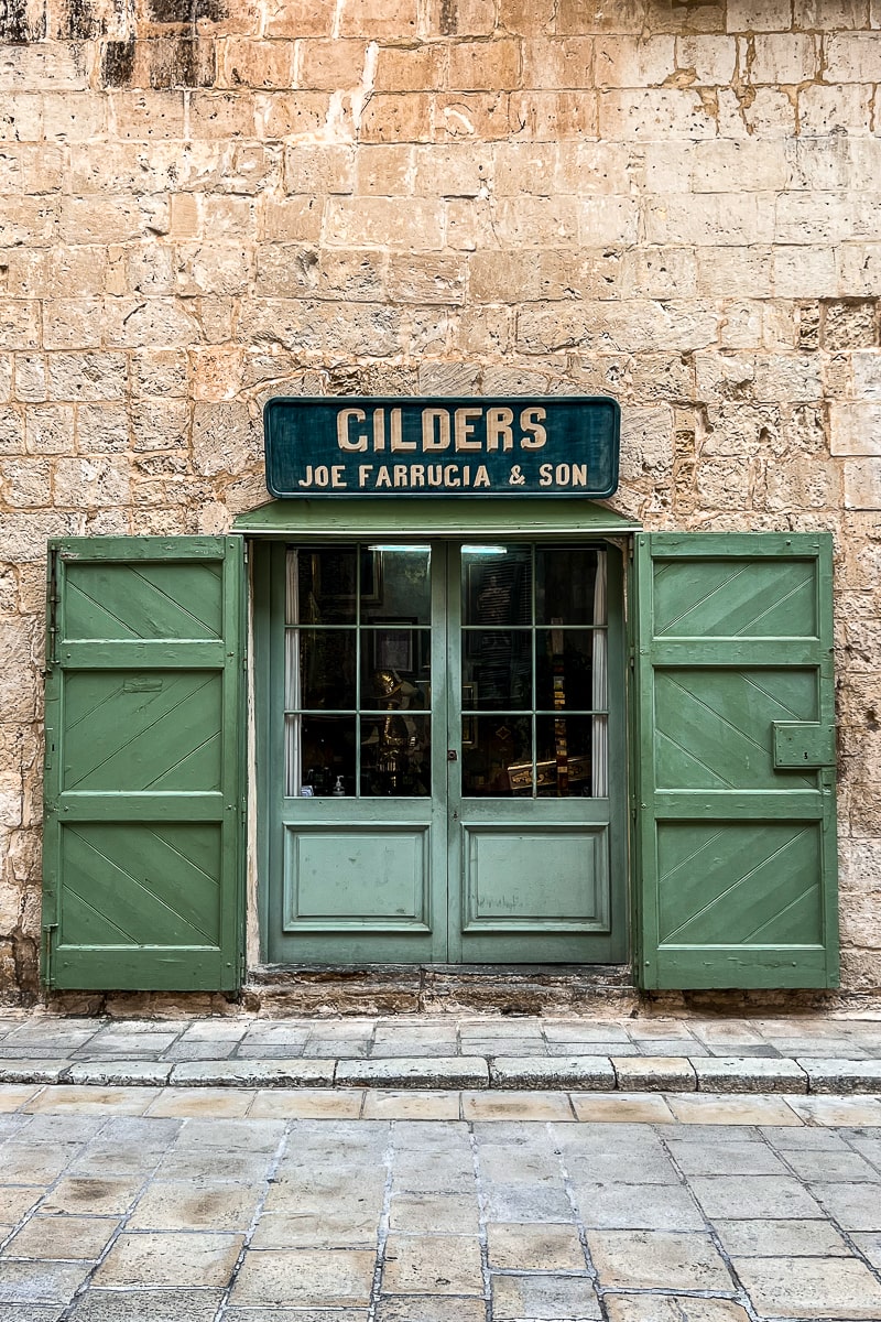 A sand colored wall with a small entrance to a shop made of green wooden doors that are swung open