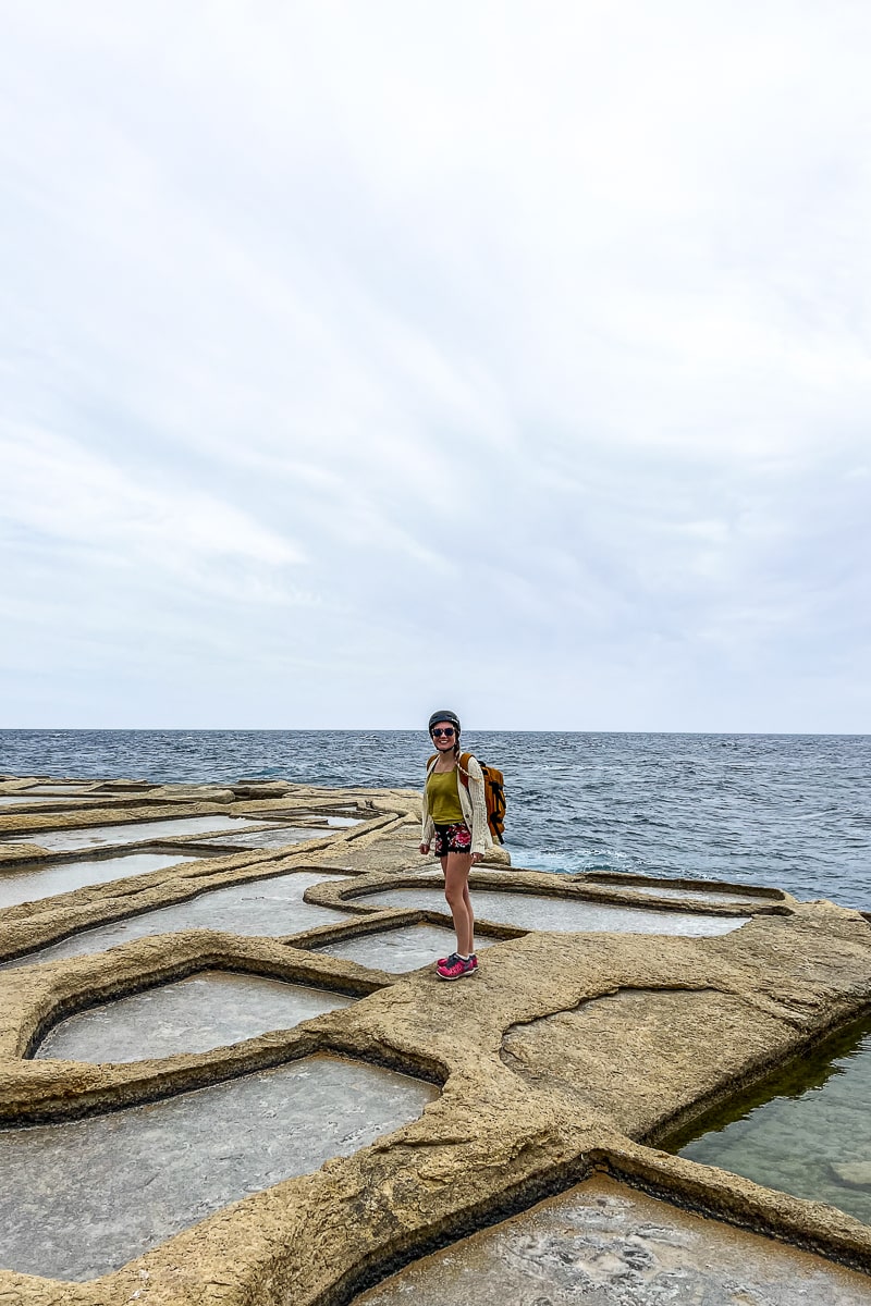 A woman stood among man-made salt pans holding shallow sea water and the sea behind