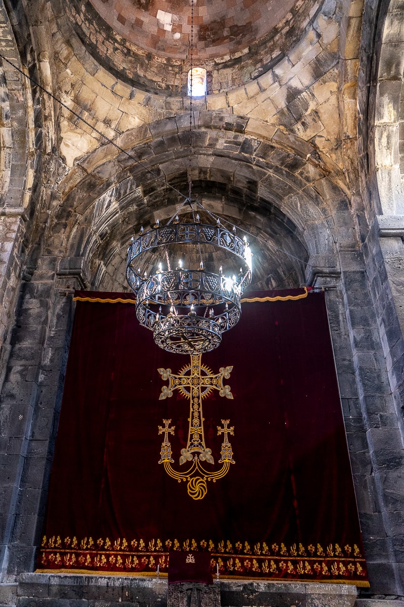 Inside a stone church, a chandelier hanging with a red and gold cloth depicting crosses behind