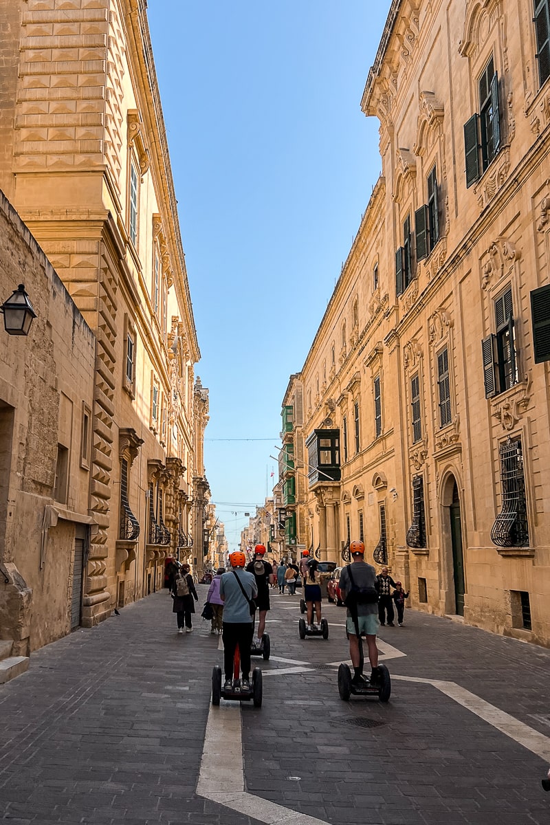 A group of people Segwaying down a street with tall sandy colored buildings on either side