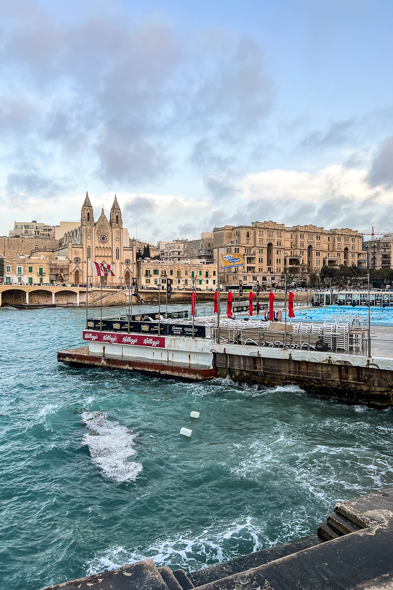 A rough sea and outdoor pool with sandy colored buildings behind including a double spired church