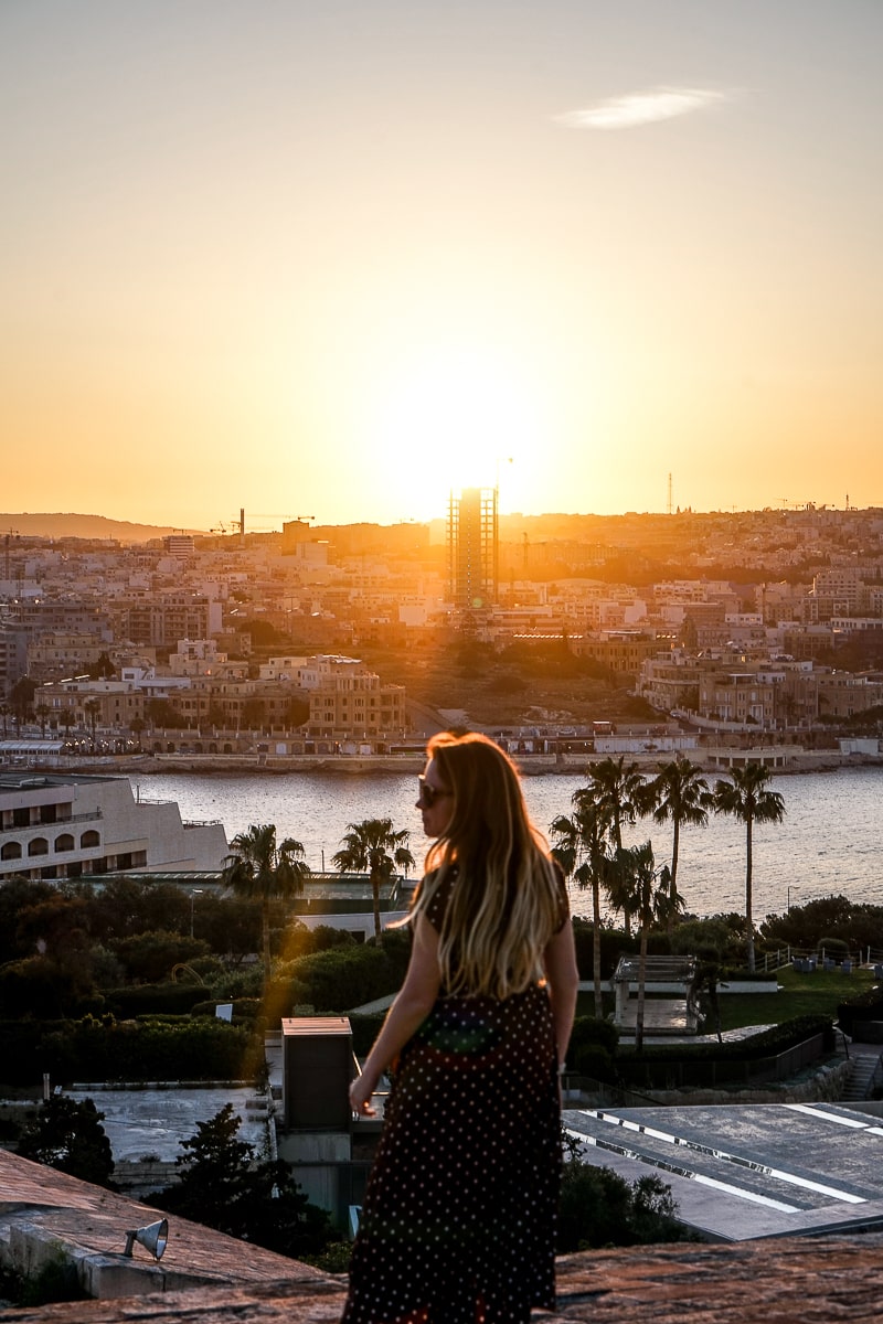 A woman on a wall with a town, half-built skyscraper, and the sun setting behind