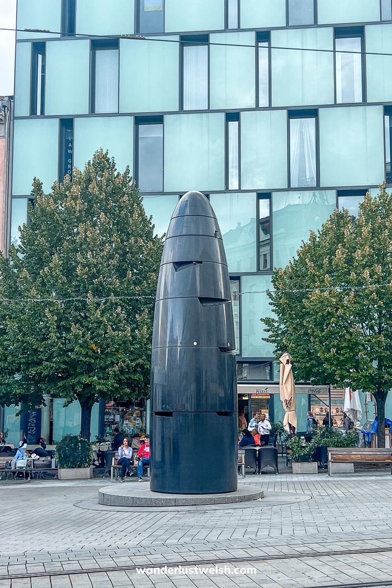 A large black statue shaped like an upright bullet in a city sqaure