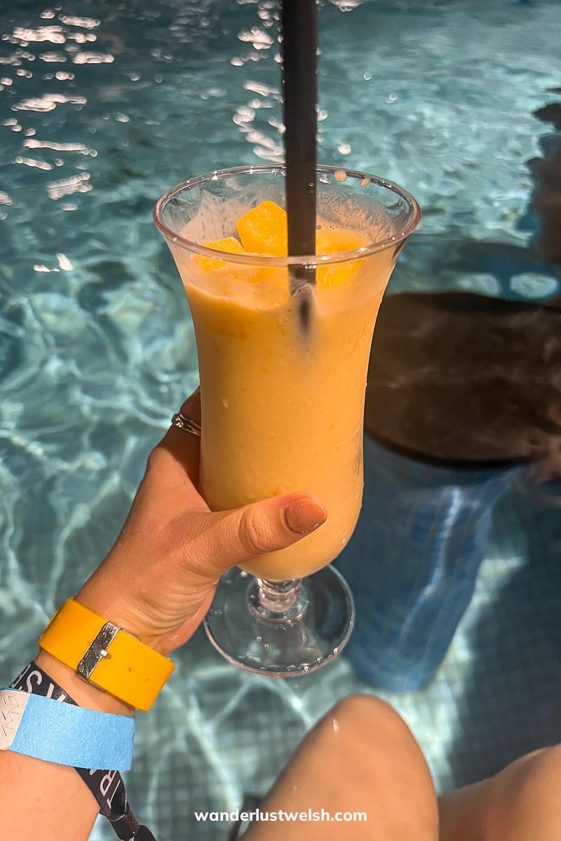 Someone holding a tropical smoothie in a tall glass in front of a spa pool