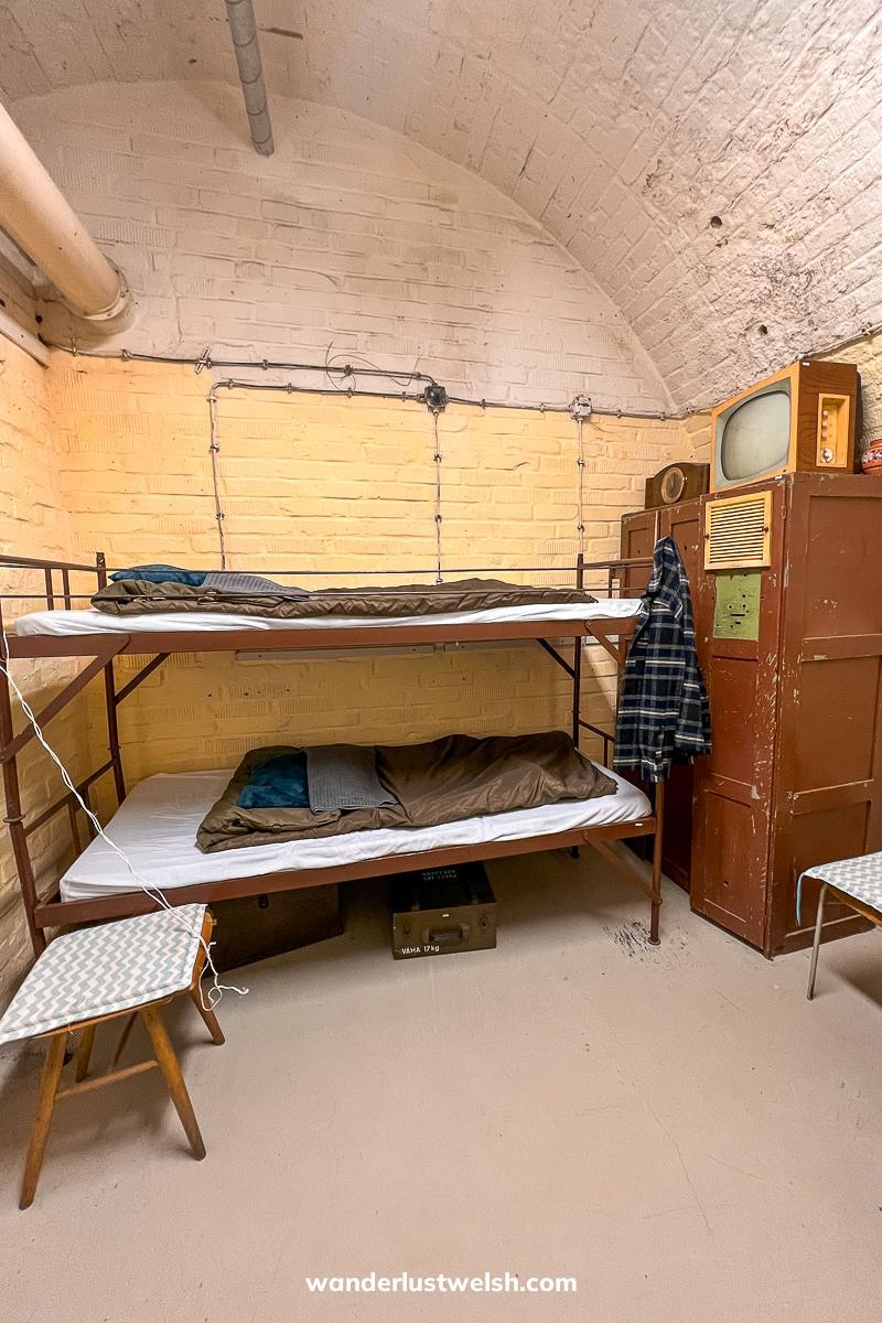 A cold war bunker bedroom with cold war era items like simple bunkbeds, sleeping bags, cupboards and old TV