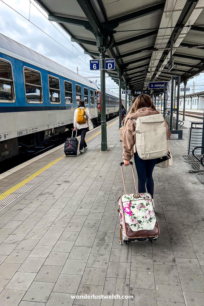 A woman dragging a flowery suitcase along a train platform next to a blue and white train in Brno station