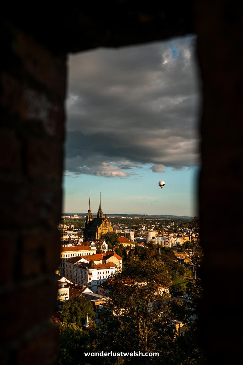 A view through an arrowslit of Brno - Double spired cathedral of St Peter and Paul and a hot air balloon at dusk