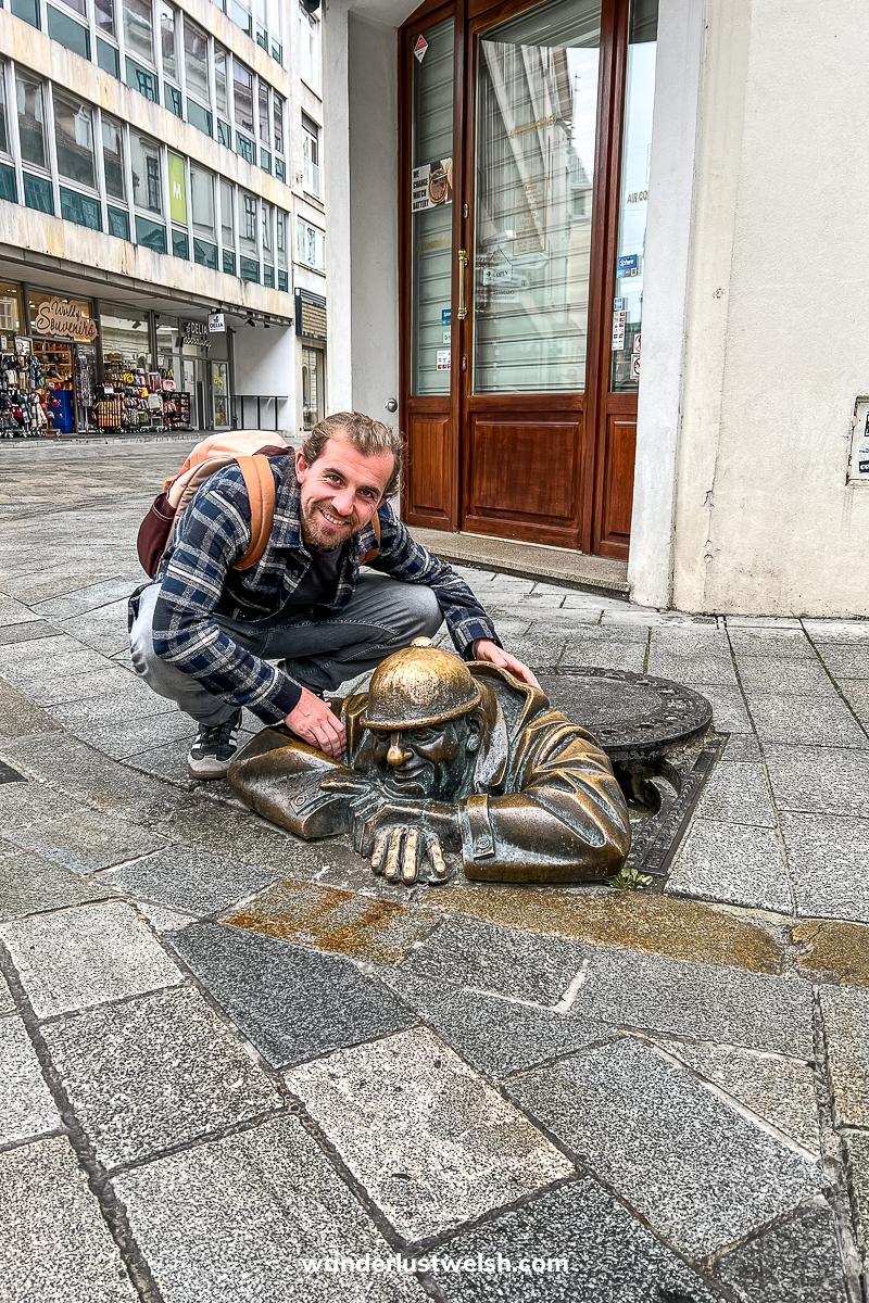 A man crouching next to a bronze sculpture of a man climbing out of a manhole cover