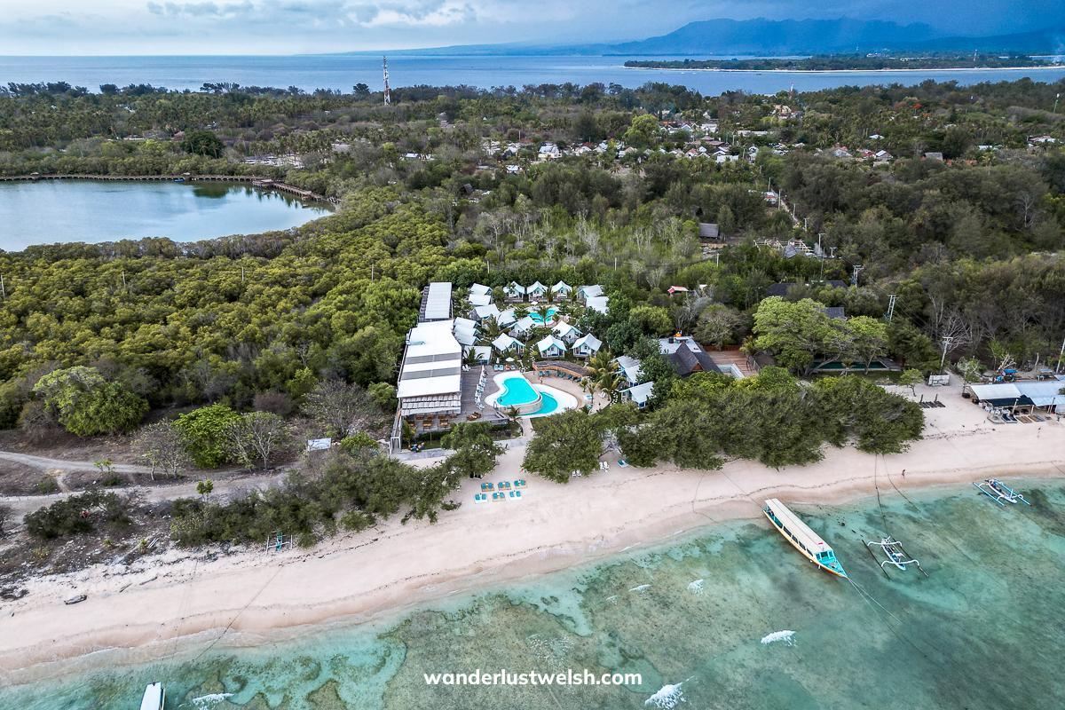 Aerial shot of Le Pirate Meno hotel with shacks and a bright blue pool, the beach, sea and an inland lake surrounded by trees