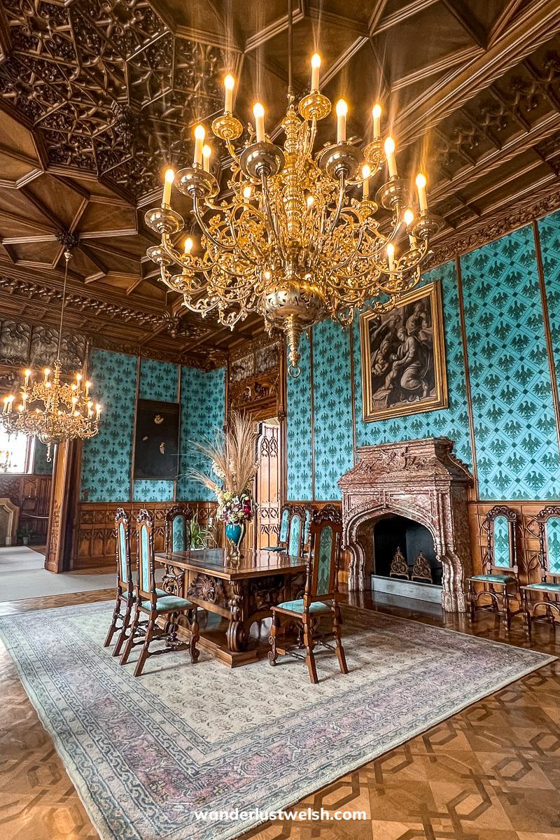 A lavishly decorated room with chandeliers, wooden carved ceiling, dark wooden dining furniture and blue/teal wallpaper