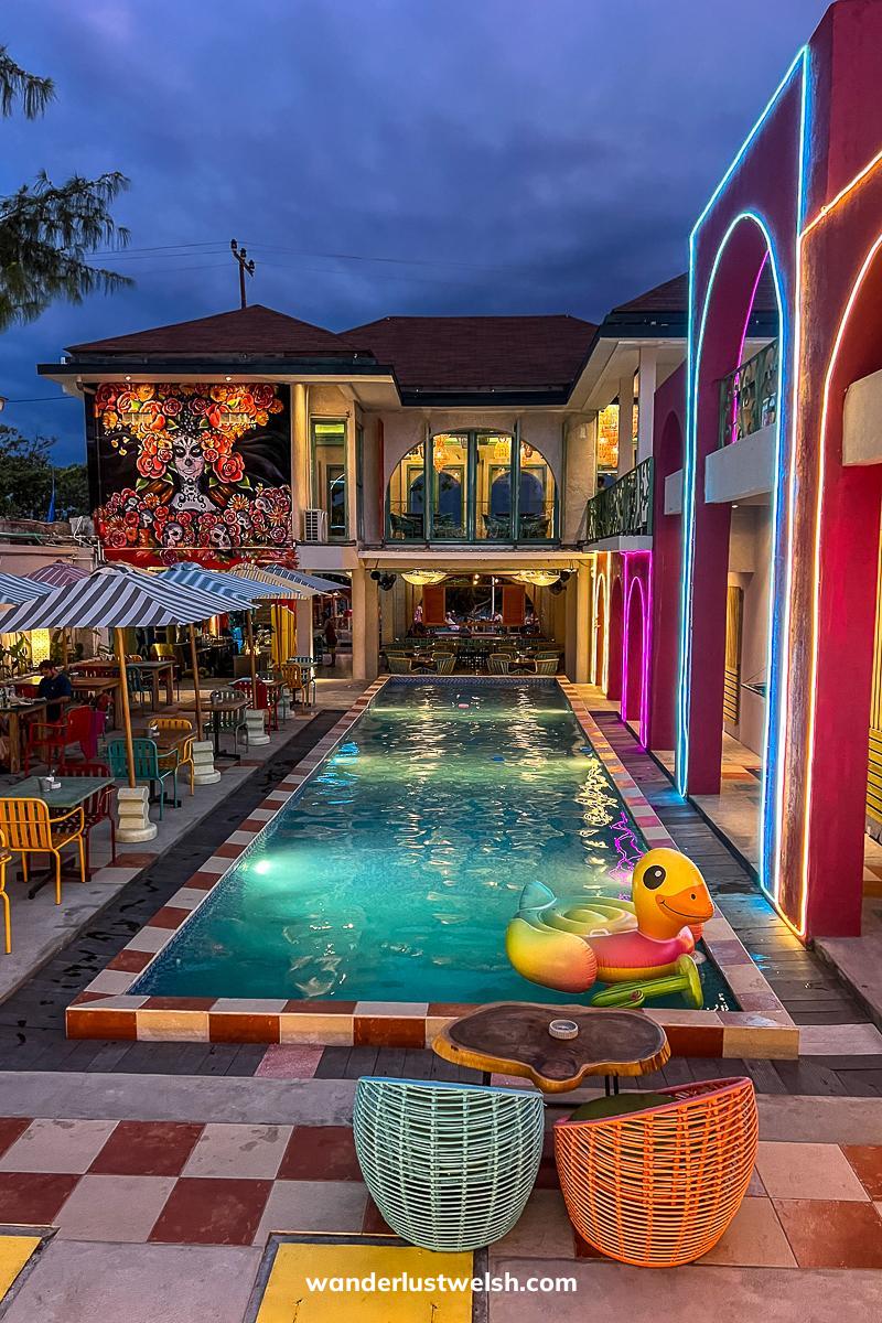 A pool at a Mexican restaurant at night - the pool is lit with lights and neon colors and has Mexican street art and a large rubber duck
