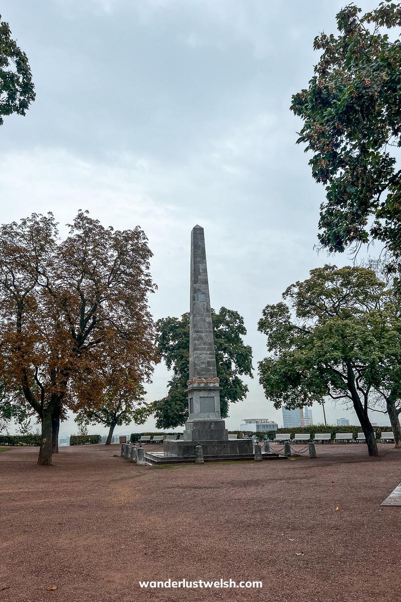 A small obelisk in a flat gravel space dotted with trees