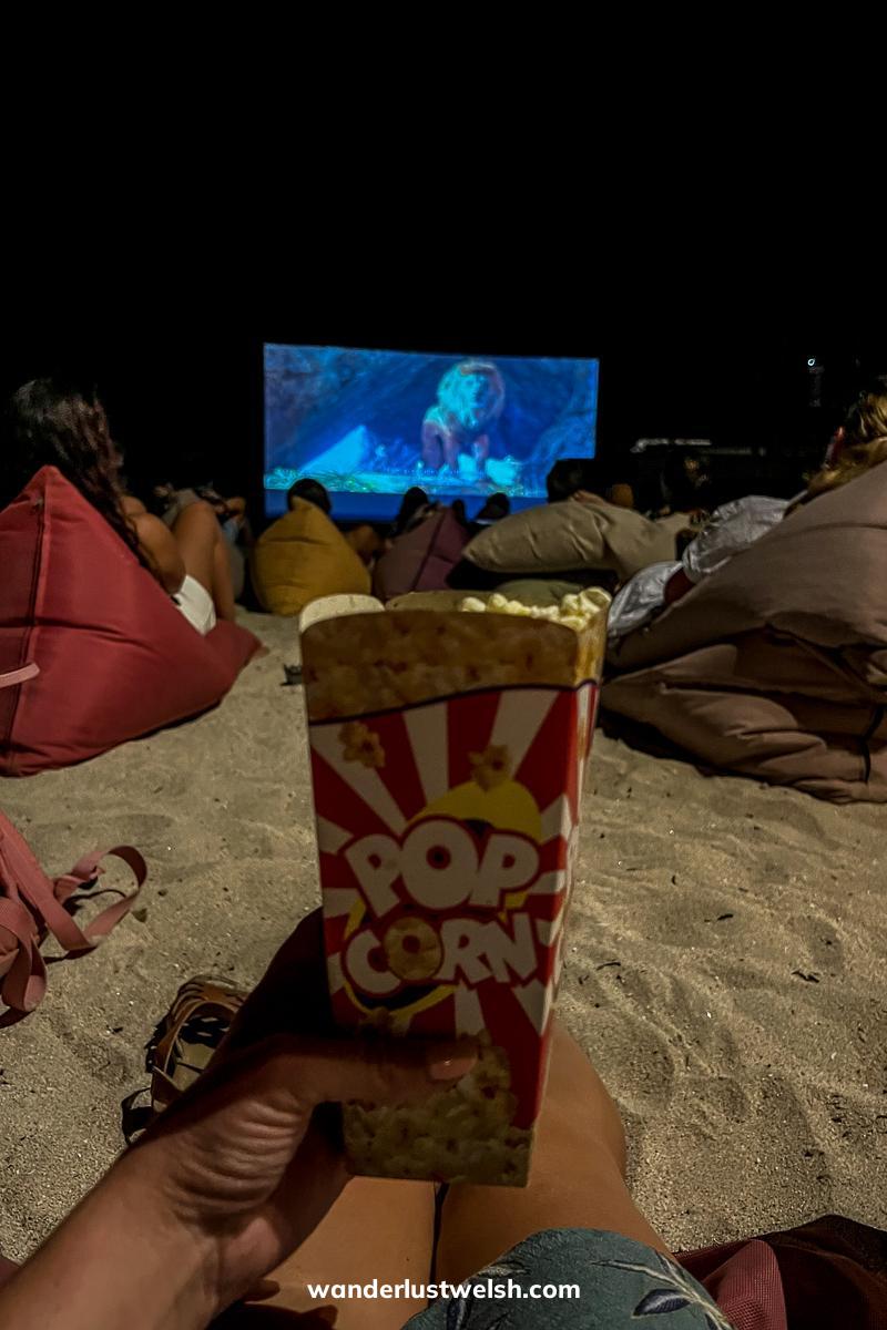 Someone holding a tall box of popcorn with a large outdoor cinema screen behind and people sitting on beanbags on the beach