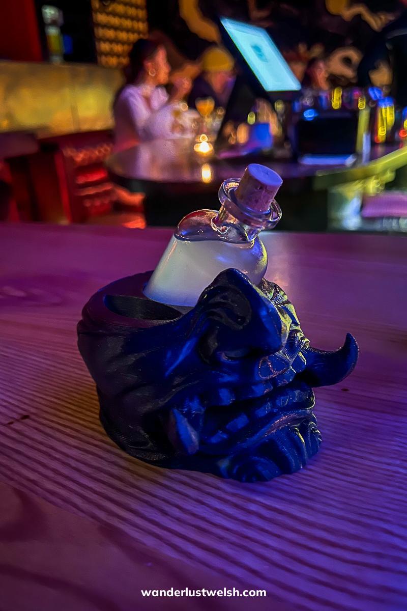 A cocktail in a small bottle that looks like a potion sitting on a demonic face