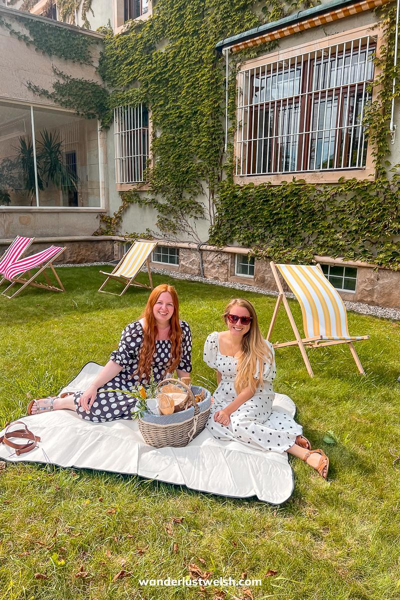 Two women enjoying a picnic on a blanket on a lawn in front of a villa