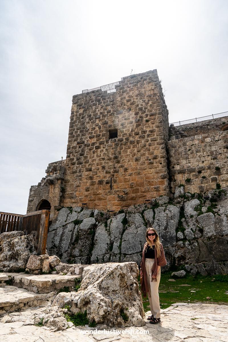 A woman standing in front of a part of Ajloun Castle, a large stone castle on a rocky outcrop