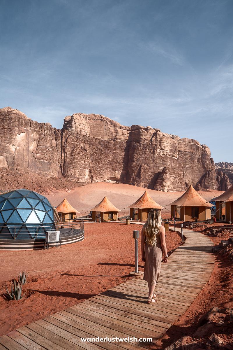 A woman walking on a boardwalk through a red sand desert with glass domes and tents