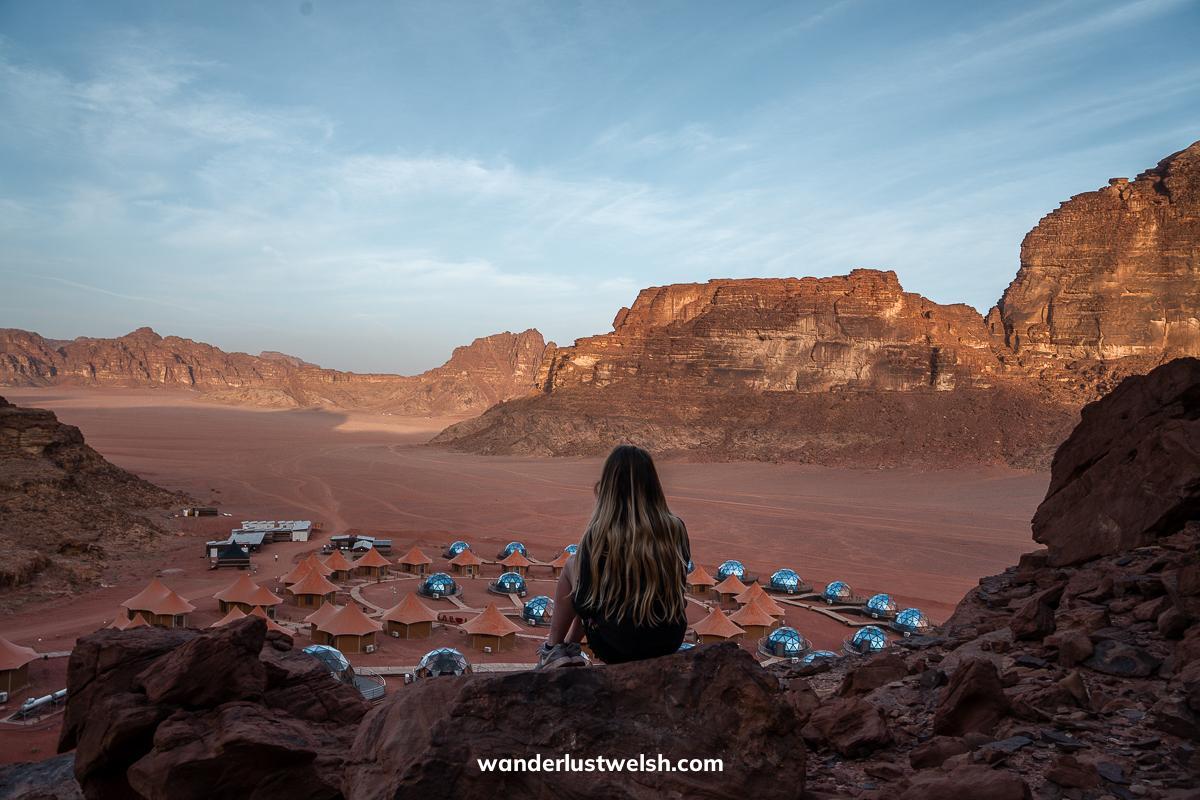 A woman sitting on a rocky cliff looking over Memories Aicha - a resort in Wadi Rum with glass domes