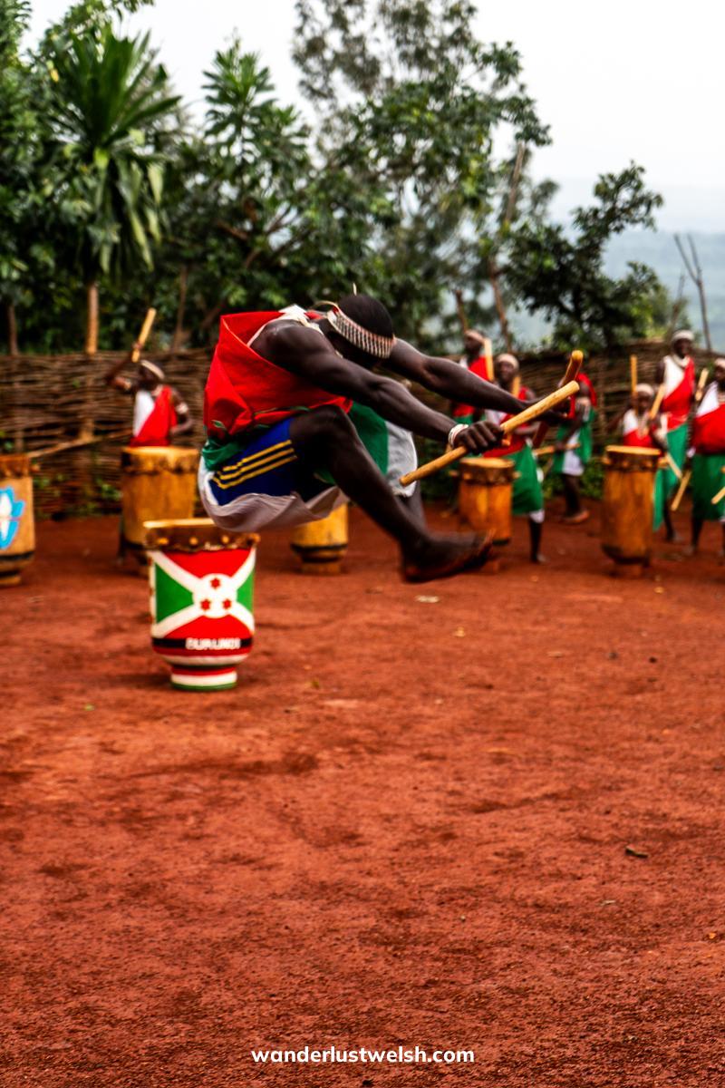 A man in tribal Burundi outfit jumping high and tucking his knees, surrounded by drummers