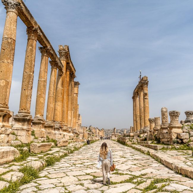 A woman walking along a wide Roman road with large columns on either side