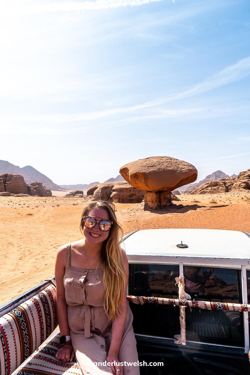 A woman sitting in the back of a truck in the desert of Wadi Rum and a mushroom shaped rock in the background